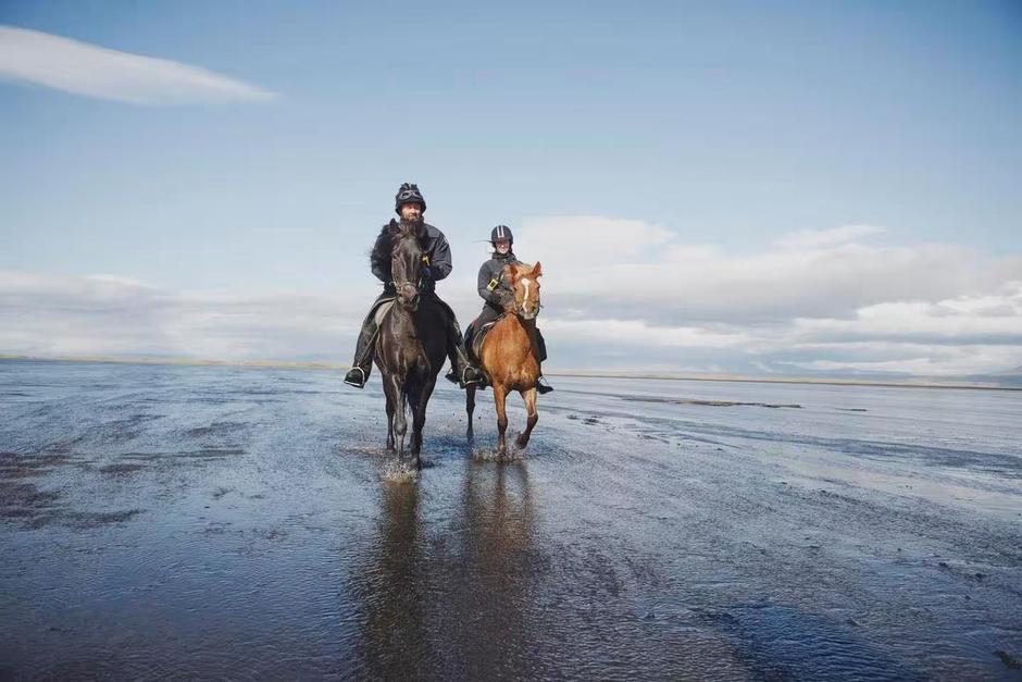 A couple on horseback galloping along a black sandy beach